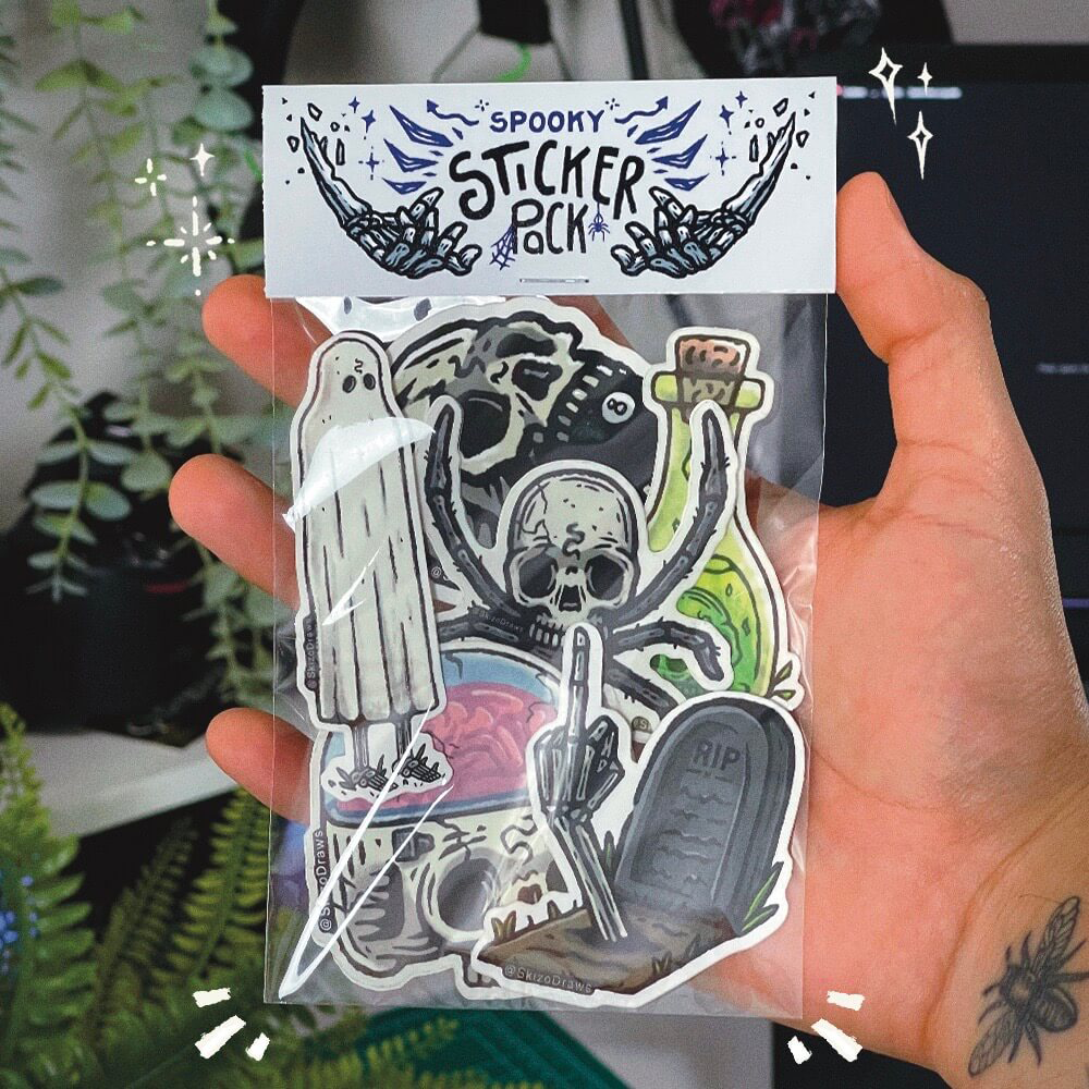 Mão a segurar um pack de stickers com desenhos de temática assustadora e um cabeçalho a dizer «SPOOKY STICKER PACK», com plantas ao fundo.