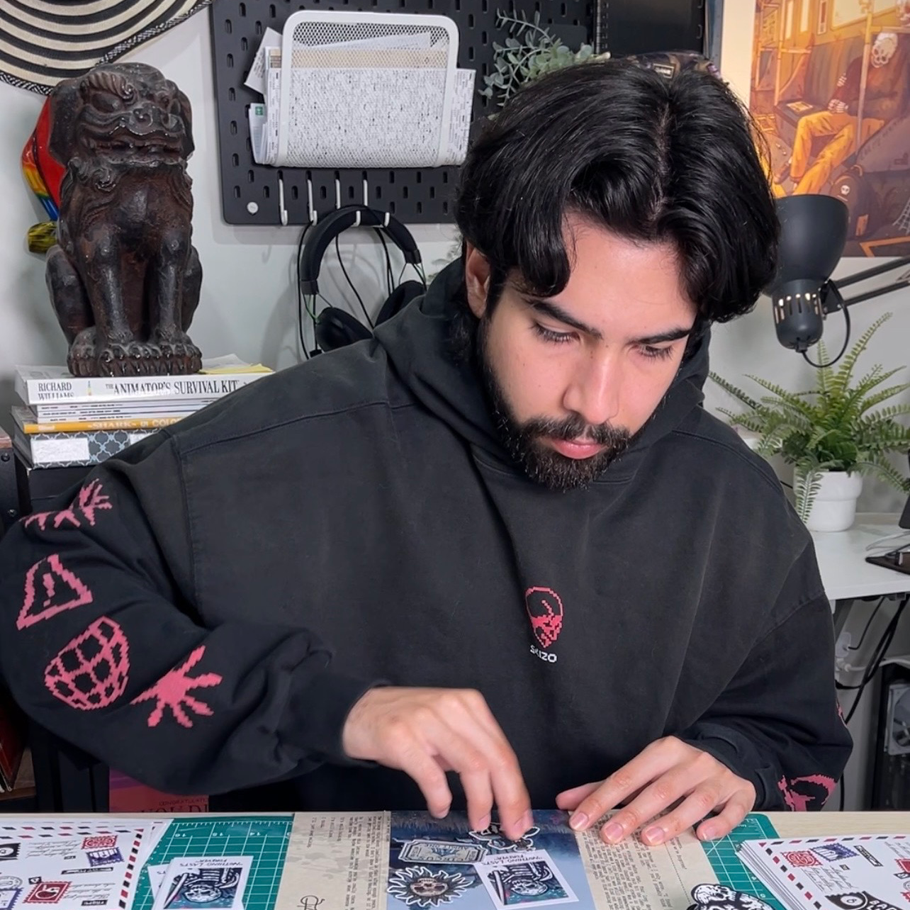 Um homem de camisola preta com capuz sentado no seu estúdio a organizar autocolantes na mesa à sua frente, com esculturas e obras de arte ao fundo.