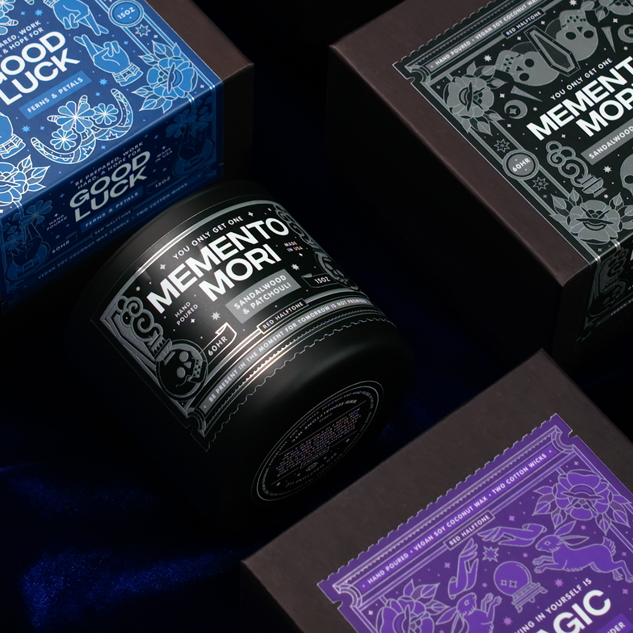 Decorative candle boxes with intricate designs and silver thin labels on roll, including "Memento Mori" and "Good Luck,".
