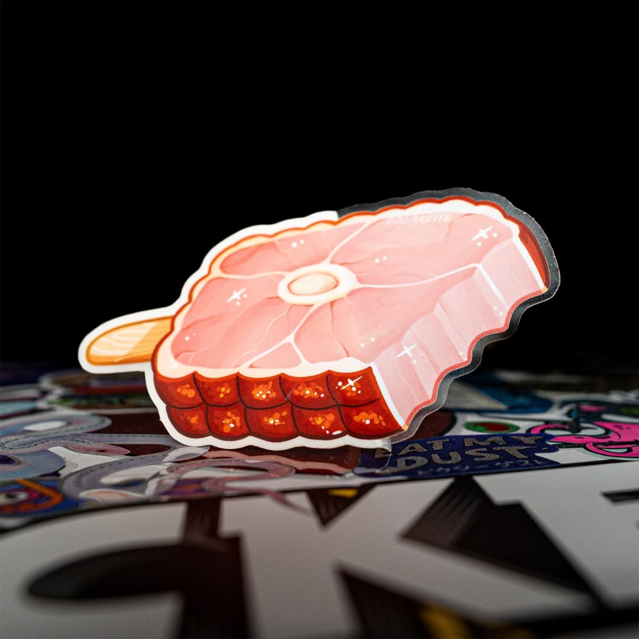 A piece of ham formed as a popsicle, with a wooden handle and colorful artwork in the foreground against a black bakground.