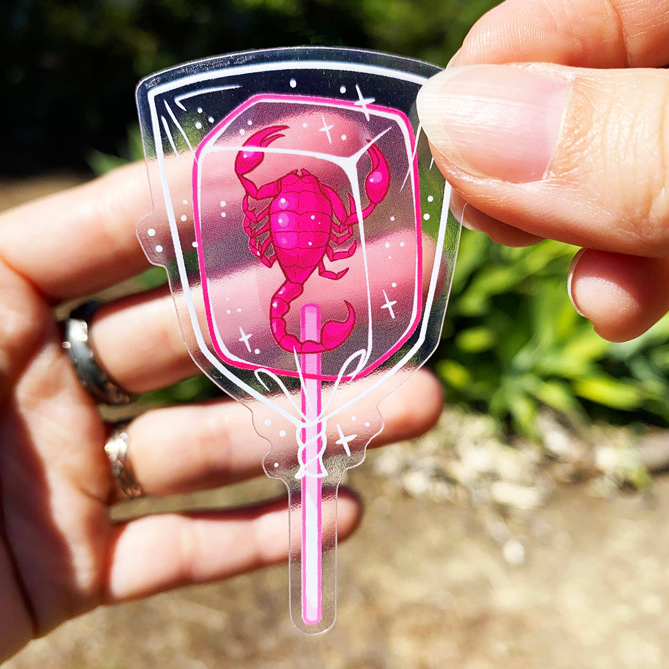 Hand holding a transparent sticker of a pink scorpion inside a lollipop design.