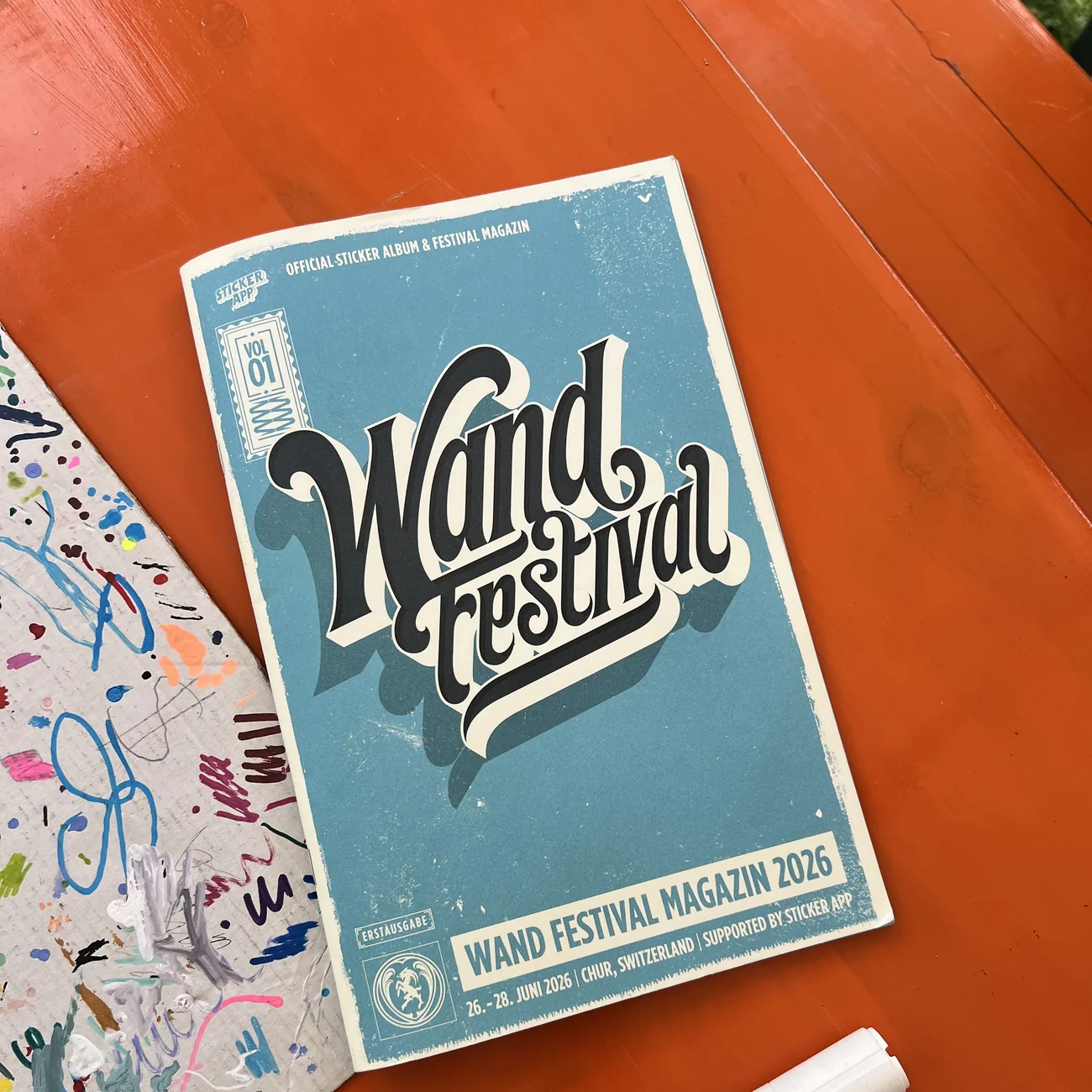 Caderneta oficial de autocolantes e revista do Wand Festival 2026, com o apoio da StickerApp, ao lado de um marcador de tinta numa mesa laranja