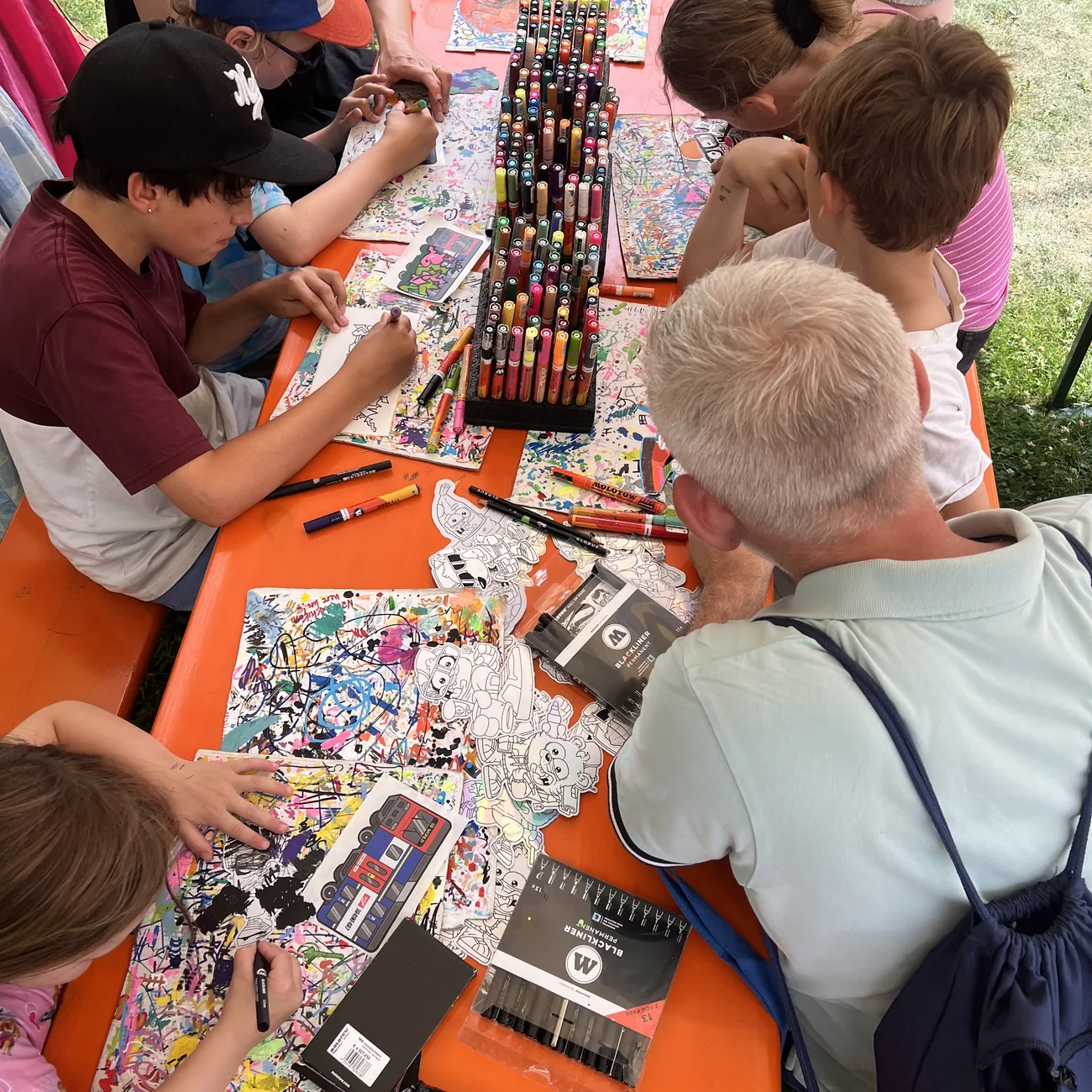 Crianças a pintar autocolantes recortados Brainfart à volta de um suporte de marcadores de tinta num workshop de festival ao ar livre