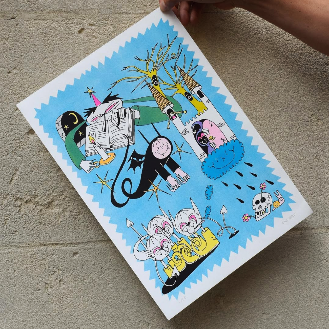 Hand holding a print with cartoon characters, skulls, a castle, and others on a light blur background, set against a rugged concrete wall.