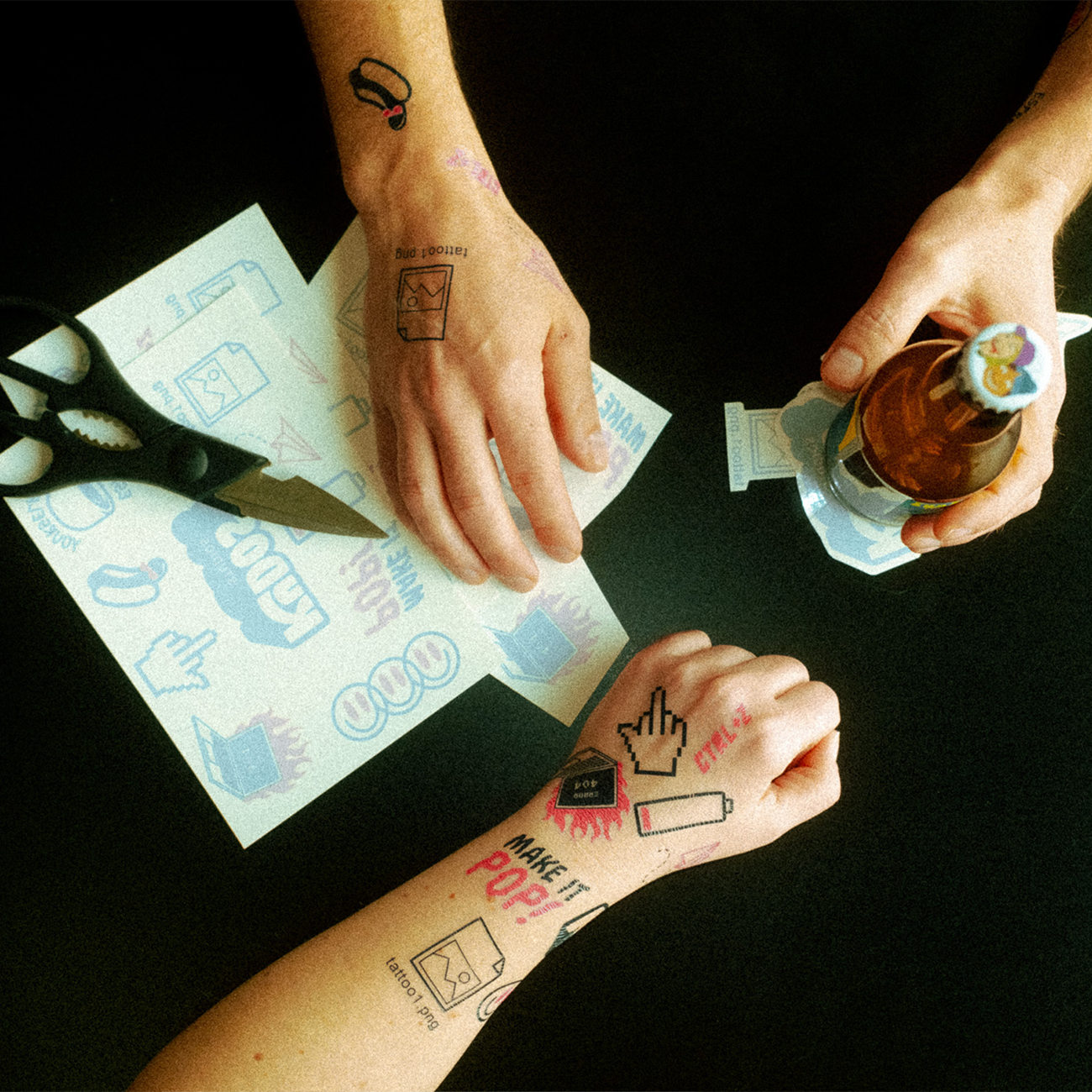 Three hands displaying colorful temporary tattoos, with scissors and a sheet of uncut tattoos on a dark surface.