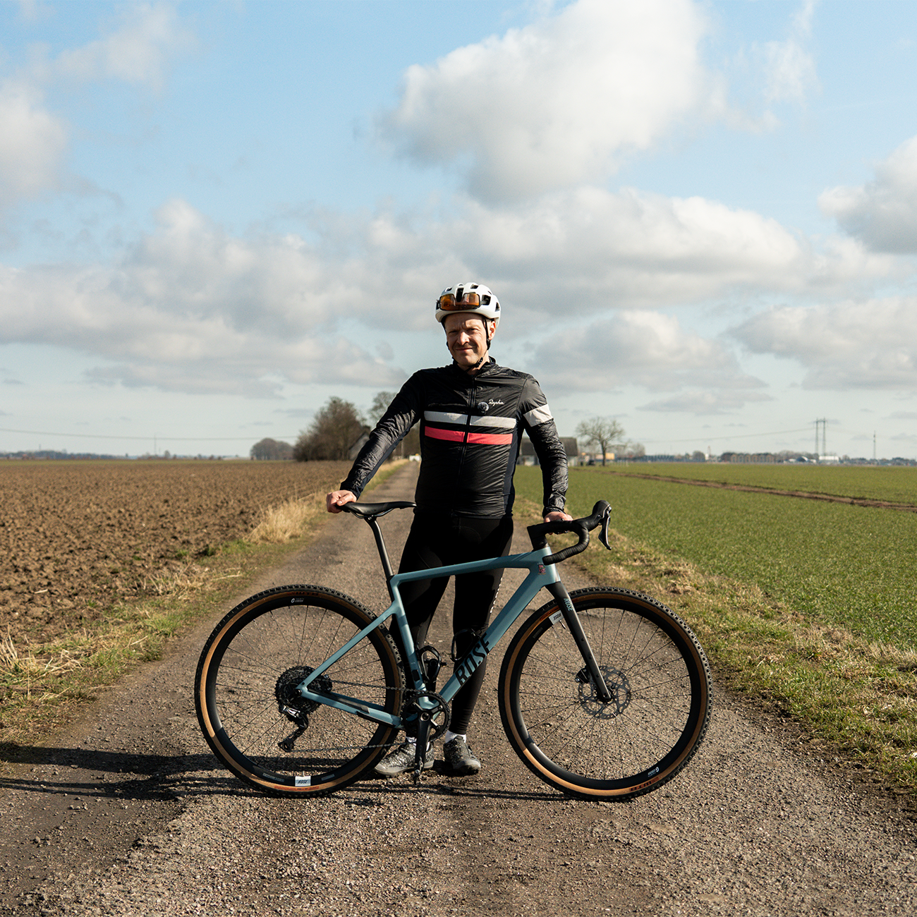 Person in cycling gear standing on a gravel road holding a bike, surrounded by open fields under a partly cloudy sky.