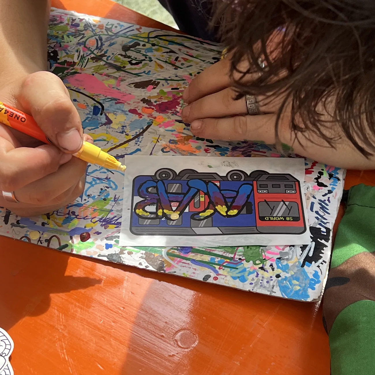 Uma mão pinta uma etiqueta do comboio S8 World com um marcador de tinta amarelo no workshop Brainfart x StickerApp