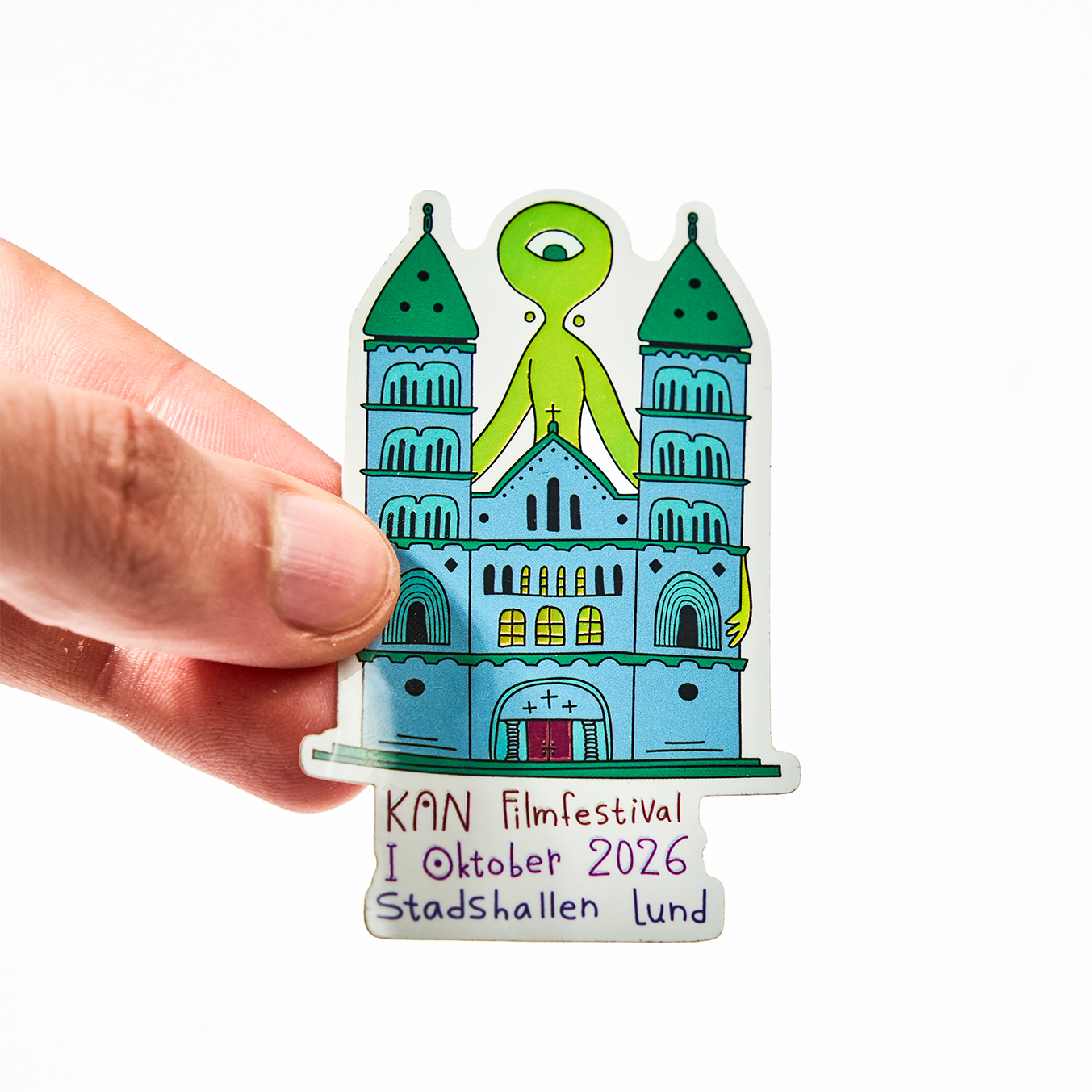 Mão a segurar um autocolante recortado para o KAN Filmfestival, outubro de 2026 no Stadshallen de Lund, com uma catedral e um extraterrestre verde de um só olho
