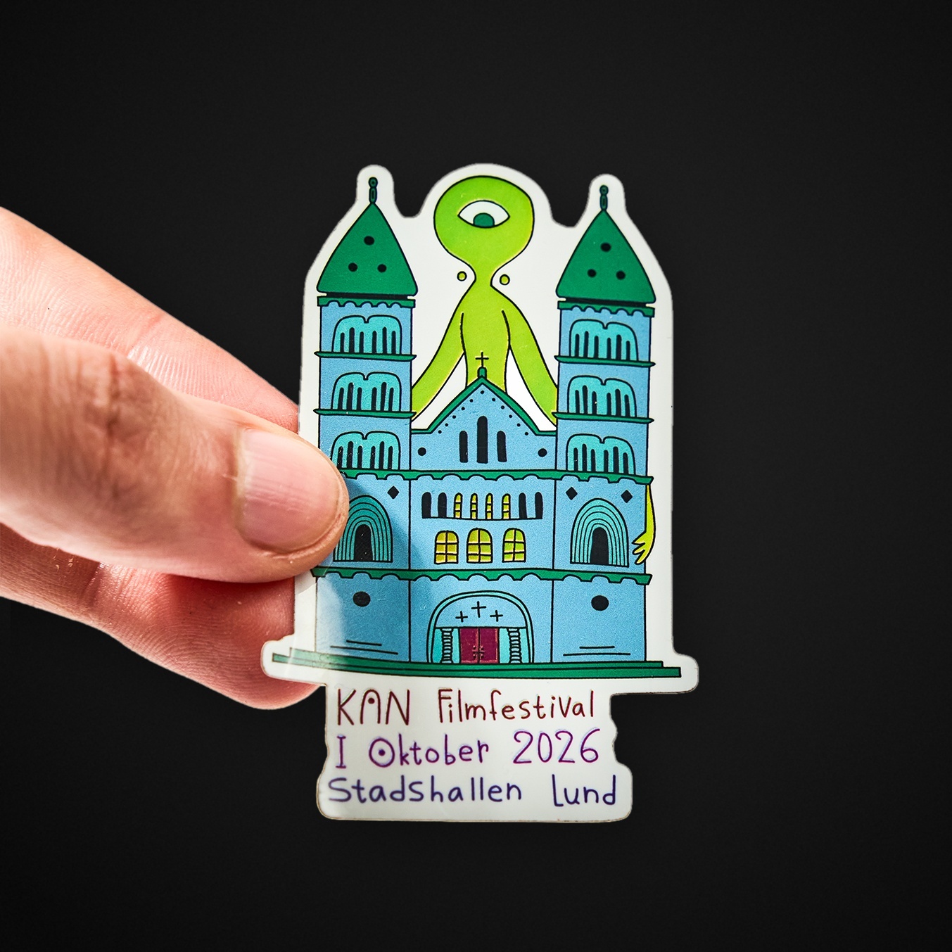 Mão a segurar um autocolante recortado de uma catedral com uma criatura verde de um só olho e o texto “KAN Filmfestival I Oktober 2026 Stadshallen Lund”
