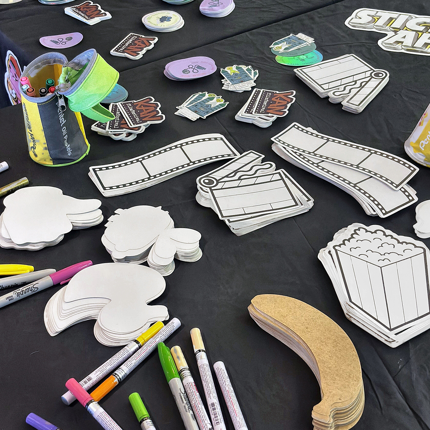 Mesa para criar autocolantes no KAN Film Festival, com autocolantes recortados em branco em forma de pipocas, fitas de filme e claquetes, autocolantes de papel kraft e marcadores de tinta