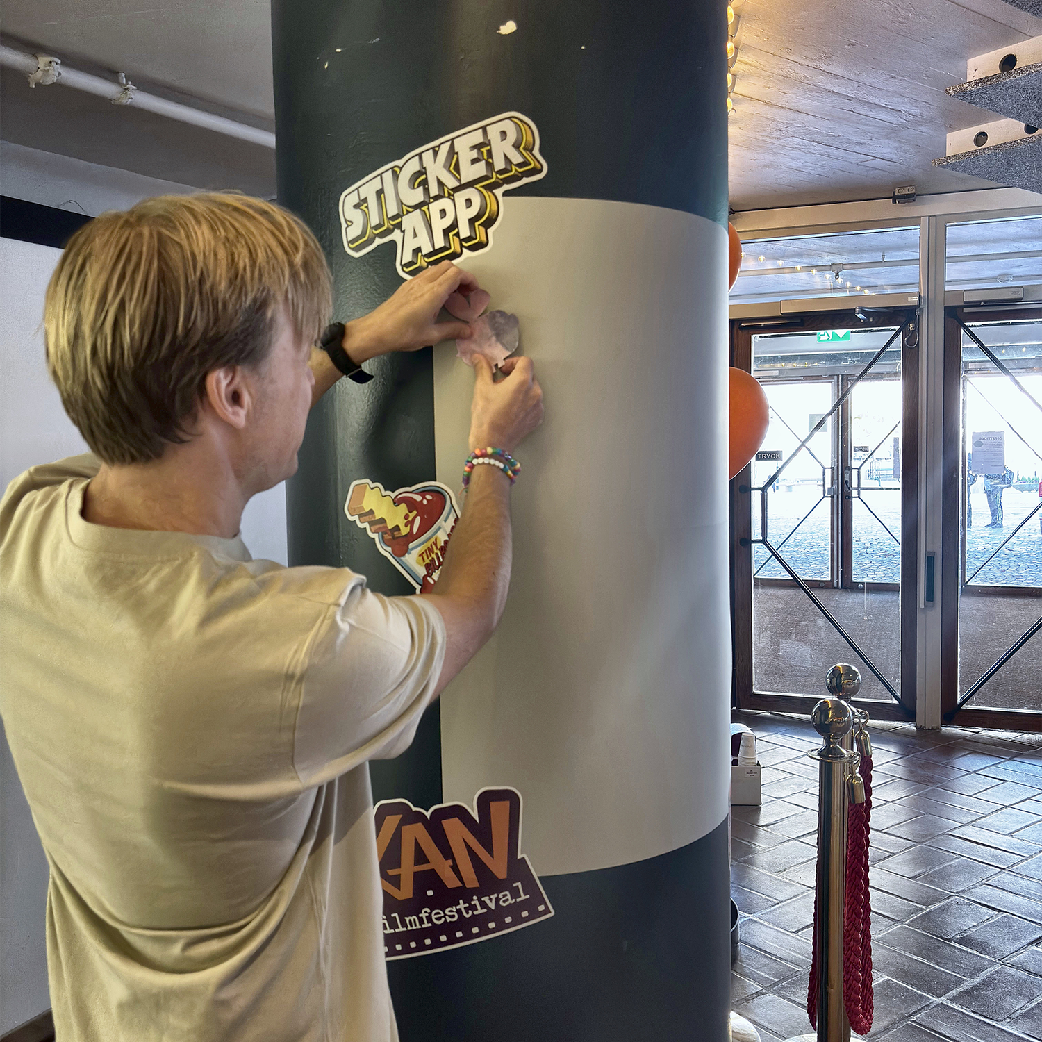 Homem a colar um autocolante numa parede de autocolantes da StickerApp à volta de um pilar no KAN Film Festival