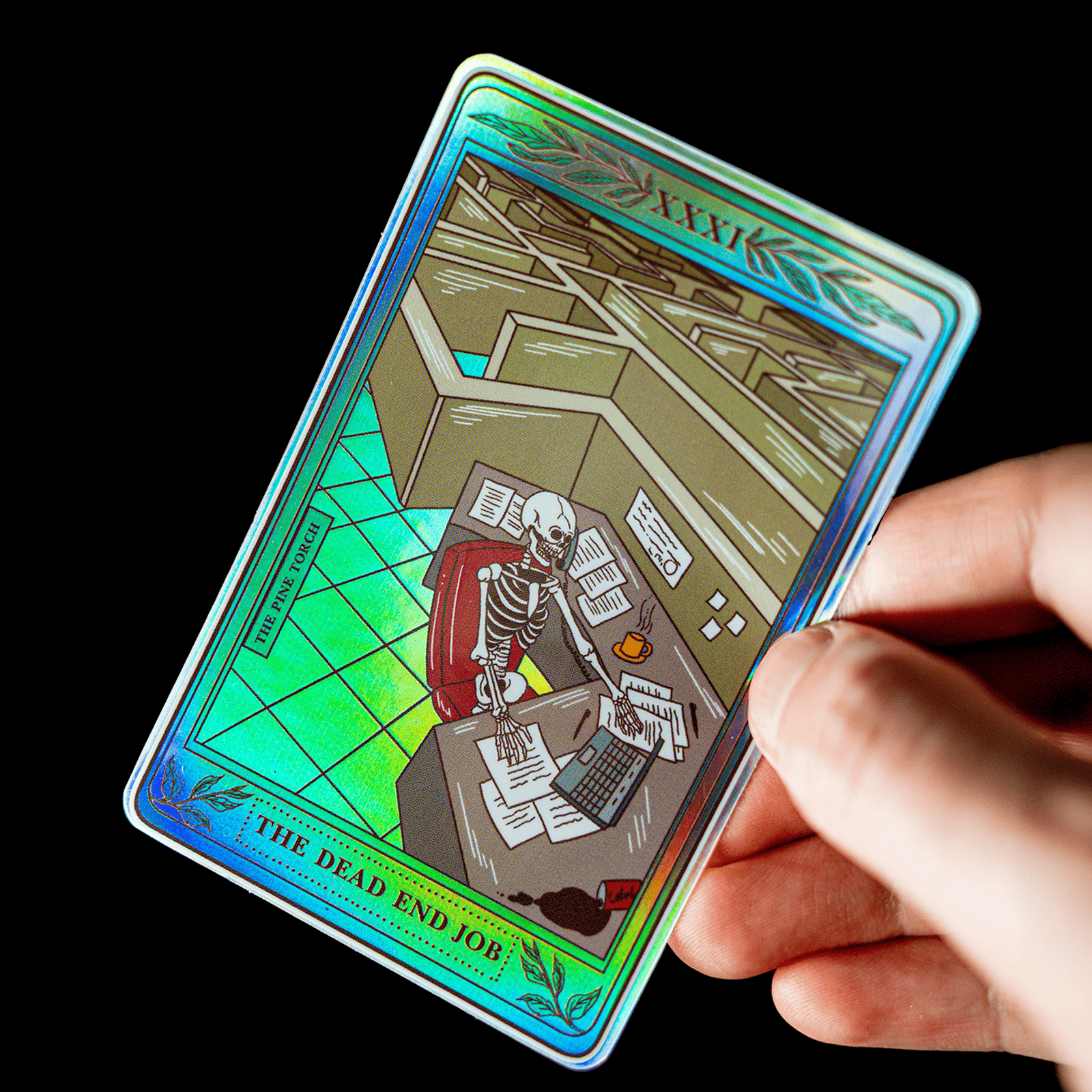 Mão a segurar um autocolante holográfico de carta de tarot com o título "The Dead End Job" e um esqueleto sentado a uma secretária de escritório