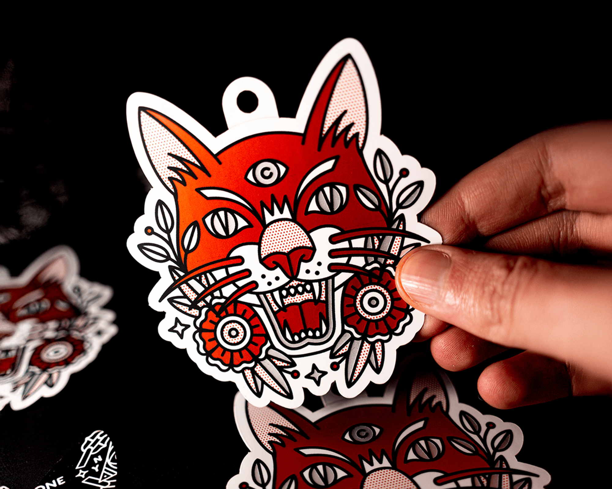 Mão segurando um sticker de etiqueta de gato vermelho estilizado com três olhos, boca aberta e decorações florais em um fundo preto.