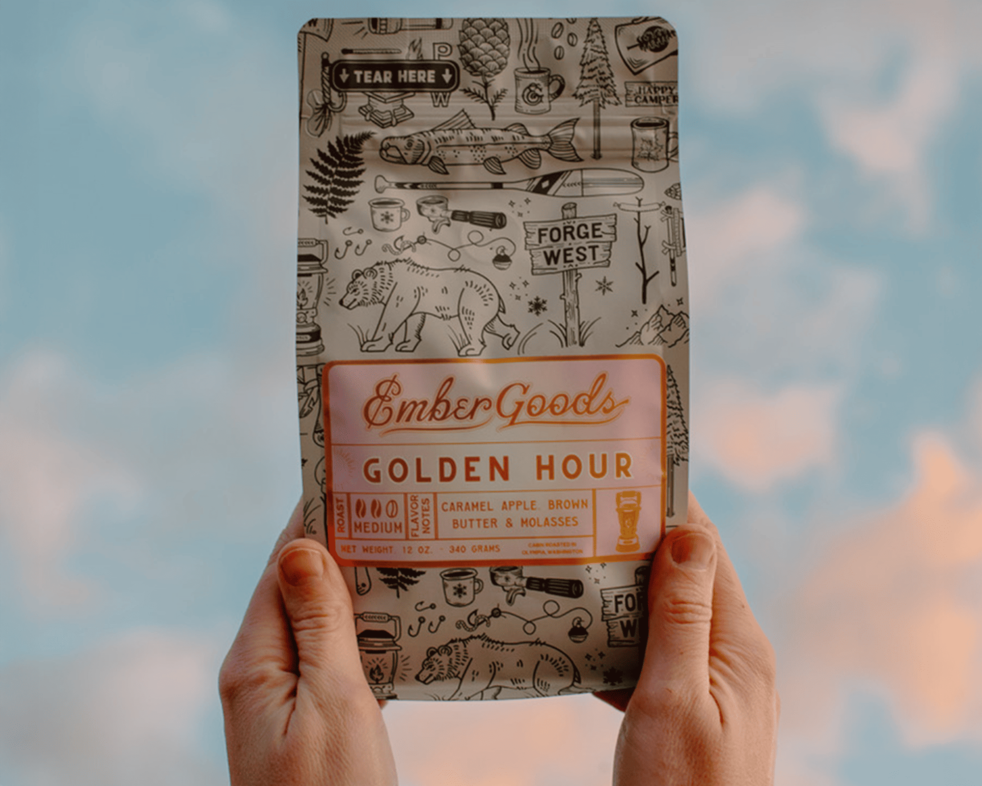 Mãos a segurar um saco de café Golden Hour da Ember Goods com uma etiqueta metalizada e o céu como fundo